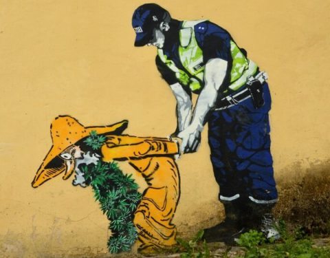 Grafite em muro - No grafite o Policial prende senhor idoso que veste capa amarela e tem sua barba feita de folhas de maconha