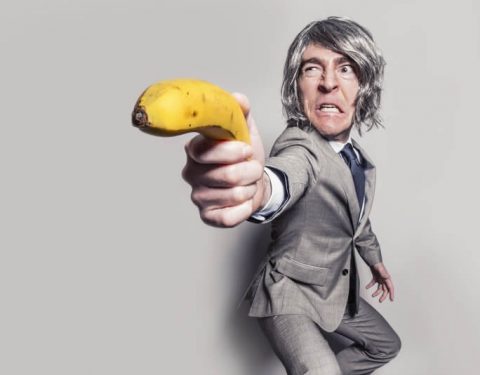 Homem em terno cinza segurando banana como se fosse uma arma representando os efeitos reais do estado de bem-estar social